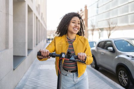 Téléchargez les photos : Joyeux jeune femme avec les cheveux bouclés en utilisant scooter électrique dans la rue et avoir du plaisir. - en image libre de droit
