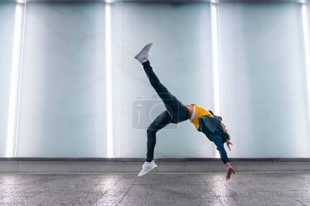 Photo for Talented young man dancing against white wall with lights and practicing break dance. - Royalty Free Image