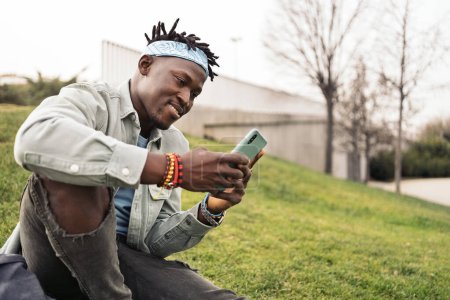 Photo for Handsome and cool african man sitting in the grass and using his cellphone. - Royalty Free Image