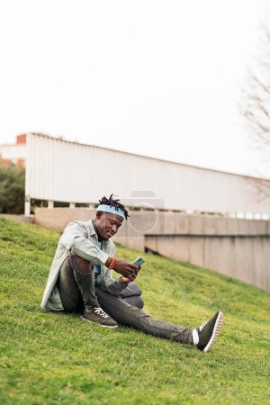 Téléchargez les photos : Beau et cool homme africain assis dans l'herbe et en utilisant son téléphone portable. - en image libre de droit
