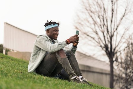 Téléchargez les photos : Beau et cool homme africain assis dans l'herbe et en utilisant son téléphone portable. - en image libre de droit