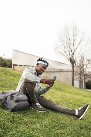 Téléchargez les photos : Heureux et cool homme africain assis dans l'herbe et en utilisant son téléphone portable. - en image libre de droit
