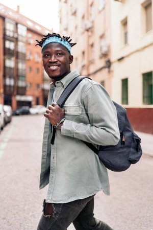 Téléchargez les photos : Attrayant garçon africain avec dreadlocks et portant un sac à dos marchant dans la ville et regardant devant. - en image libre de droit