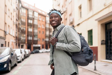 Téléchargez les photos : Attrayant garçon africain avec dreadlocks et portant un sac à dos marchant dans la ville et regardant devant. - en image libre de droit