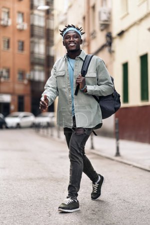 Foto de Atractivo chico africano con rastas y mochila caminando por la calle y mirando al frente. - Imagen libre de derechos