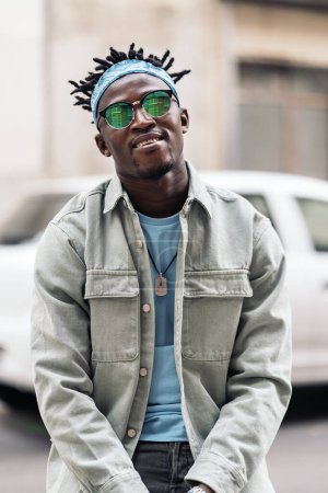 Téléchargez les photos : Jeune homme africain confiant avec des lunettes de soleil debout dans la rue et regardant la caméra. - en image libre de droit