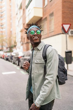 Téléchargez les photos : Attrayant garçon africain avec dreadlocks et lunettes de soleil marchant dans la ville et regardant devant. - en image libre de droit