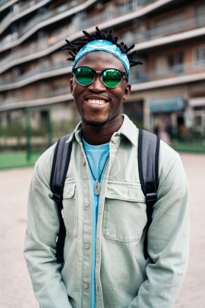 Foto de Chico afro fresco con gafas de sol sonriendo y mirando a la cámara en la calle. - Imagen libre de derechos