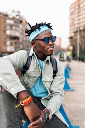 Téléchargez les photos : Jeune homme africain confiant assis dans la rue et regardant vers le côté. - en image libre de droit