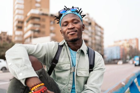 Téléchargez les photos : Jeune homme africain confiant assis dans la rue et regardant la caméra. - en image libre de droit
