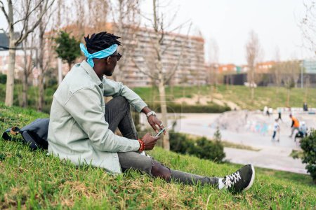 Téléchargez les photos : Cool homme africain assis dans l'herbe et en utilisant son téléphone portable. - en image libre de droit