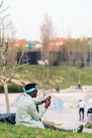 Foto de Cool joven africano sentado en la hierba y usando su teléfono móvil. - Imagen libre de derechos