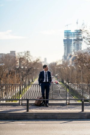 Téléchargez les photos : Homme entrepreneur en costume formel debout avec les mains dans les poches sur le pont en ville et détournant les yeux - en image libre de droit