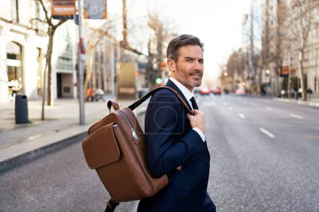 Photo for Side view of serious middle aged male entrepreneur in suit and with backpack walking across road while commuting to work - Royalty Free Image
