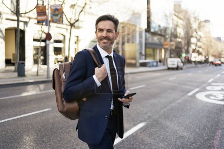 Photo for Middle aged male entrepreneur in formal suit browsing cellphone while walking in city and commuting to work - Royalty Free Image