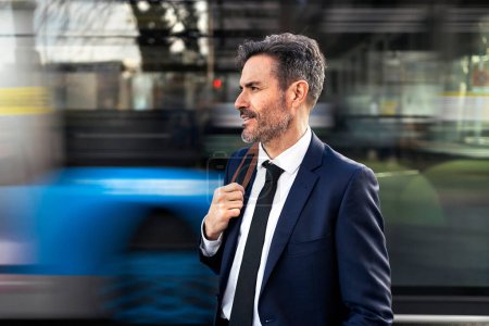 Photo for Mature male entrepreneur in suit standing against blurred moving transport in street while commuting to work - Royalty Free Image