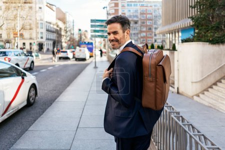 Téléchargez les photos : Vue arrière de l'entrepreneur masculin mature avec sac à dos en cuir marchant le long de la rue et se rendant au travail le matin - en image libre de droit