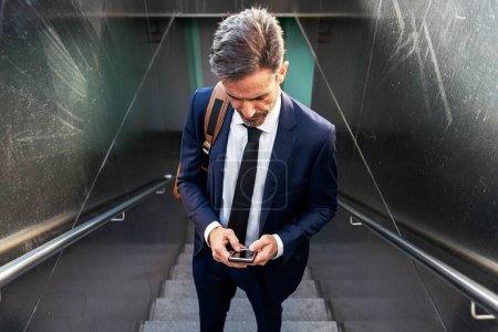 Foto de Alto ángulo de confianza empresario varón maduro en traje y con teléfono móvil de pie en la escalera de paso subterráneo y centrado - Imagen libre de derechos