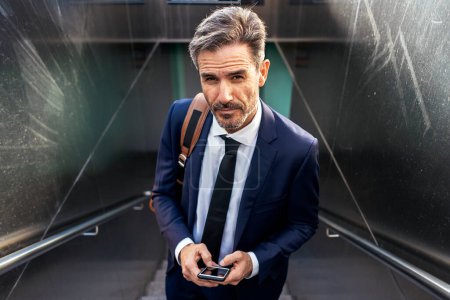 Téléchargez les photos : High angle of confident mature male entrepreneur in suit and with mobile phone standing on staircase of underpass and looking at camera - en image libre de droit