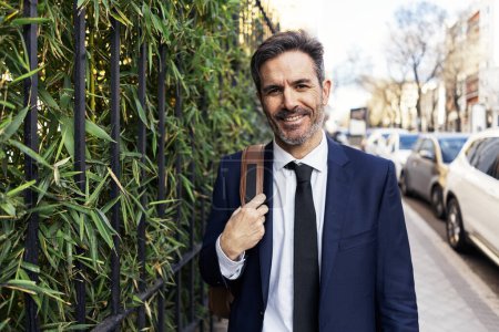 Photo for Positive middle aged businessman in suit with backpack smiling and looking at camera while standing near metal fence covered with green ivy on sunny day in city - Royalty Free Image