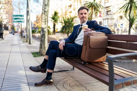 Téléchargez les photos : Charmant entrepreneur d'âge moyen en costume chic assis sur un banc avec du café pour aller en ville et détourner les yeux - en image libre de droit