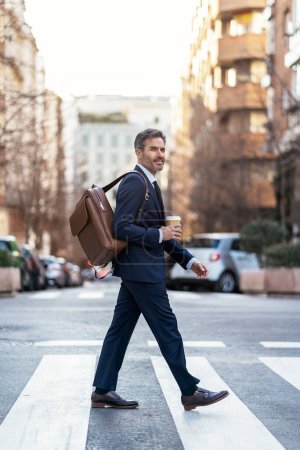 Photo for Side view of male entrepreneur in classy suit and with takeaway drink walking along crosswalk while commuting to work in morning - Royalty Free Image