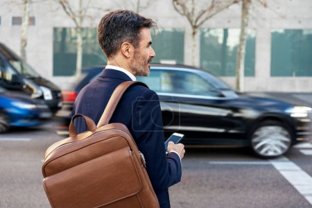 Foto de Vista trasera del empresario varón maduro con mochila de cuero caminando por la calle y viajando al trabajo por la mañana - Imagen libre de derechos