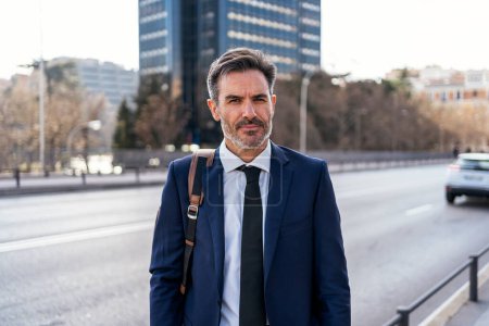 Téléchargez les photos : Positive middle aged businessman in suit with backpack smiling and looking at camera while standing on sunny day in city - en image libre de droit