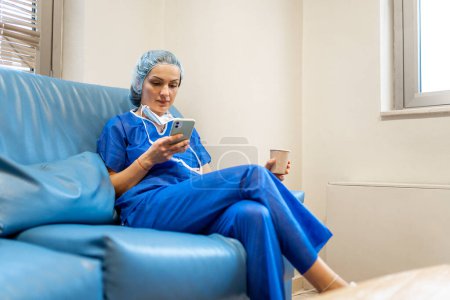 Foto de Nurse using the mobile phone during her break sitting on a sofa with a coffee - Imagen libre de derechos