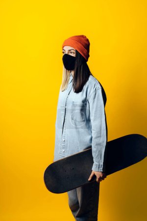 Photo for Stock photo of young expressive girl looking at camera in studio. She is holding a skateboard. She is wearing a face mask. - Royalty Free Image