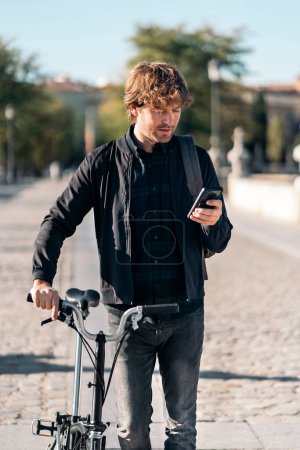 Téléchargez les photos : Photo de stock de bel homme utilisant son smartphone dans la rue et portant son vélo amovible. - en image libre de droit