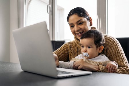Téléchargez les photos : Photo de stock de mère heureuse et son bébé regardant des dessins animés dans l'ordinateur à la maison. - en image libre de droit