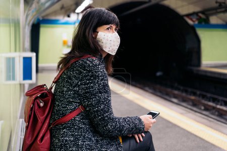 Photo for Stock photo of adult woman wearing face mask due to covid19 sitting in a bench while the metro arrives. - Royalty Free Image
