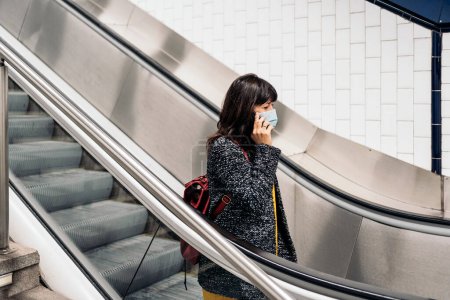 Foto de Stock photo of happy woman wearing face mask talking on the phone while standing in escalators. - Imagen libre de derechos