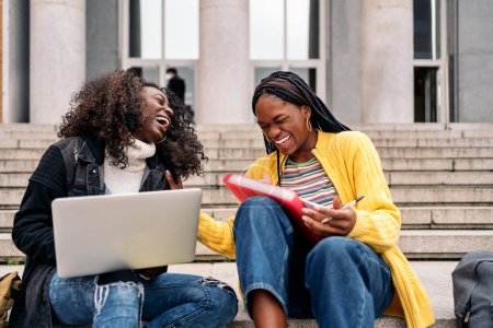 Photo for Stock photo of cheerful black friends using laptop while sitting in stairs and doing homework. - Royalty Free Image