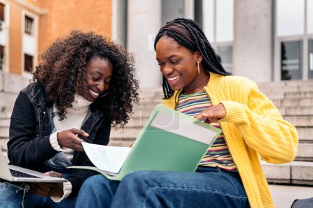 Photo for Stock photo of cheerful black friends using laptop while sitting in stairs and doing homework. - Royalty Free Image