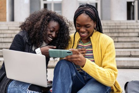 Photo for Stock photo of black cheerful friends using laptop and cellphone while sitting in stairs. - Royalty Free Image