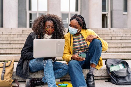 Foto de Stock photo of black friends using laptop while sitting in stairs. They are wearing face mask due to covid-19. - Imagen libre de derechos