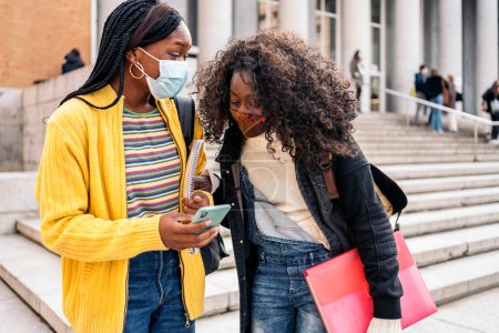 Photo for Stock photo of black female students talking and using phone. They are wearing face mask due to covid19. - Royalty Free Image