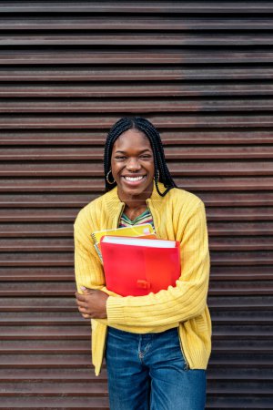 Téléchargez les photos : Photo de stock de jeune fille noire tenant un cahier, souriant et regardant la caméra. - en image libre de droit