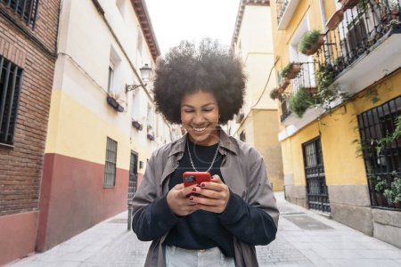 Foto de Foto de stock de hermosa mujer afroamericana sonriendo y utilizando el teléfono celular en la calle. - Imagen libre de derechos