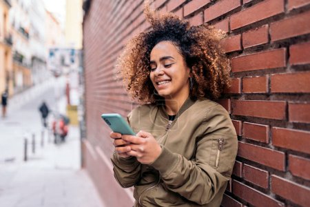 Téléchargez les photos : Photo en stock de belle femme afro-américaine souriante et utilisant un téléphone portable dans la rue. - en image libre de droit