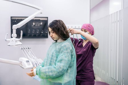 Foto de Foto de stock del dentista femenino ayudando a la paciente joven a ponerse ropa desechable para el procedimiento de cirugía. - Imagen libre de derechos