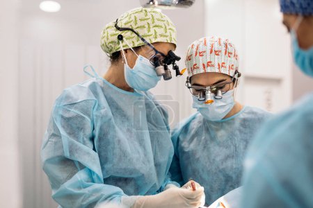 Téléchargez les photos : Photo de l'équipe de femmes portant des masques et des loupes dentaires pendant la chirurgie. - en image libre de droit