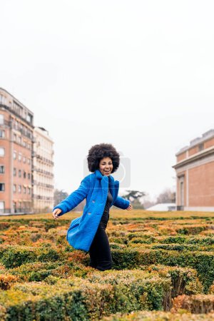 Téléchargez les photos : Photo de stock de jeune fille afro avec manteau bleu s'amuser dans un beau jardin. - en image libre de droit