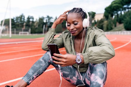 Téléchargez les photos : Photo en stock d'une sprinteuse afro-américaine assise sur la piste d'athlétisme avec ses casques, écoutant de la musique avec son téléphone portable - en image libre de droit