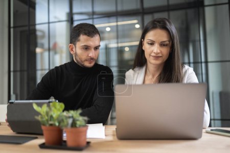 Photo for Positive young colleagues in casual clothes sitting at table with laptop and working on remote project while looking at screen of laptop. Cooperation concept - Royalty Free Image