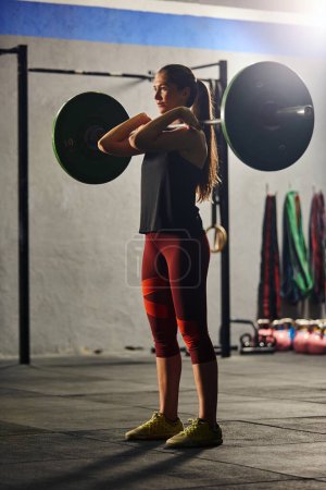 Photo for Stock photo of an adult woman in a gym standing up and lifting a barbell. She is wearing sportswear. - Royalty Free Image
