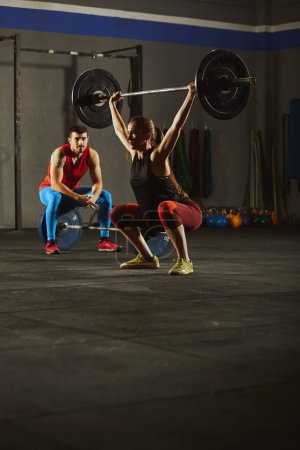 Photo for Stock photo of an adult woman squatting an lifting a barbell. There is man sitting and looking at her. They are wearing sportswear. - Royalty Free Image