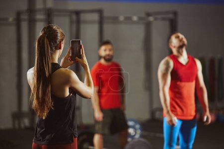 Photo for Stock photo of three adult people in a gym standing. A woman is taking a picture of two men with her smartphone. They are wearing sportswear. - Royalty Free Image